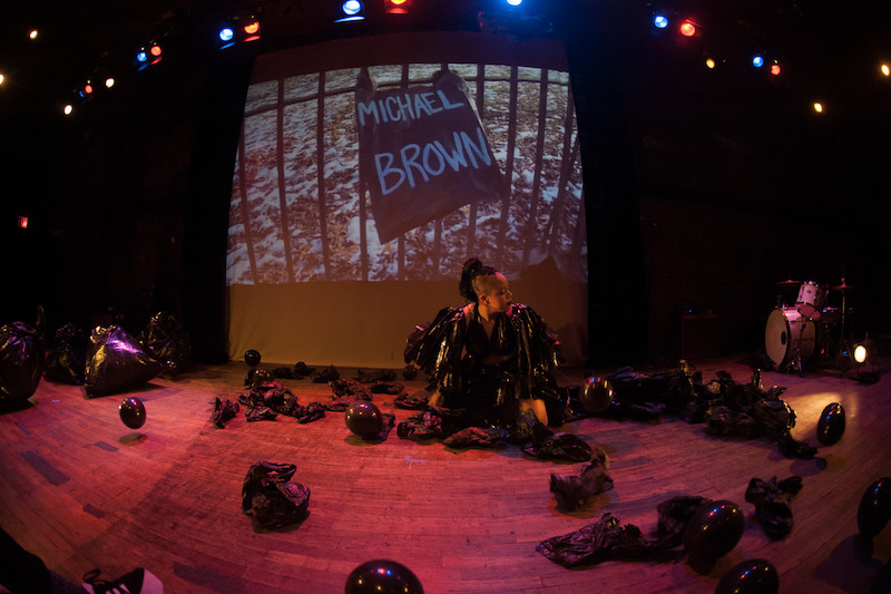 Taja Lindley in a black garbage bag costume and black garbage bags dot the stage space